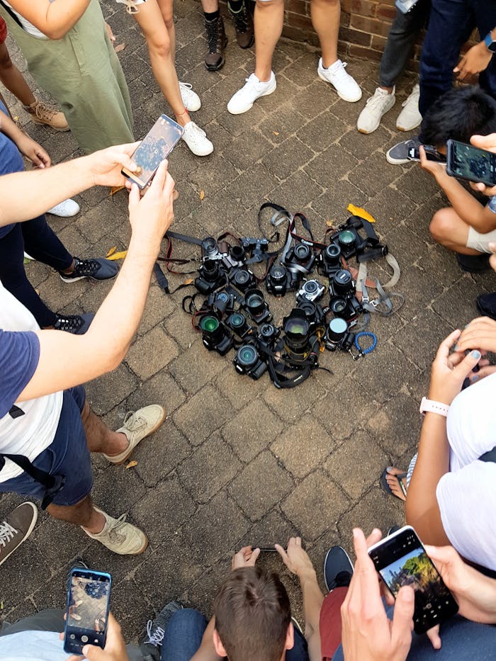 A diverse group of people using smartphones to capture a collection of cameras on the pavement.